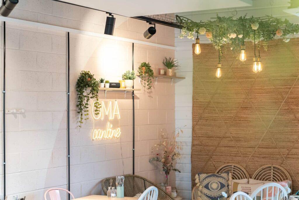 Vue d’un coin de restaurant cosy chez OMA Cantine, avec déco murale en briques peintes, néon lumineux, suspensions florales, ambiance chaleureuse et végétale.