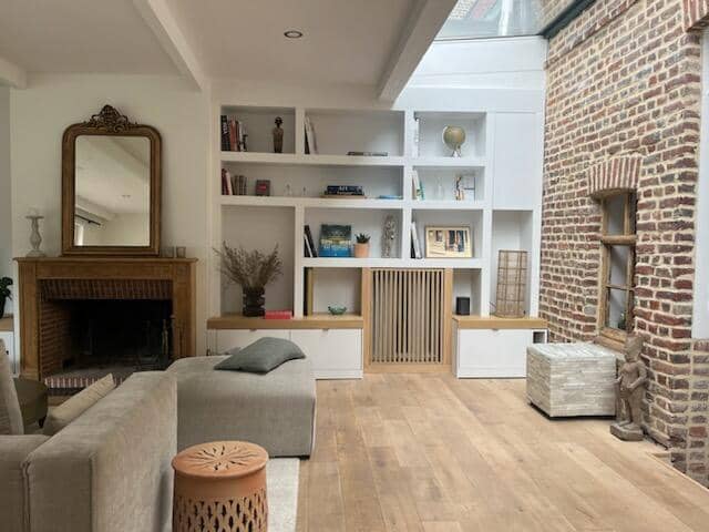 alon lumineux avec mur en briques anciennes, grande bibliothèque sur mesure blanche avec niches décoratives, parquet bois clair, et cheminée traditionnelle surmontée d’un miroir doré.