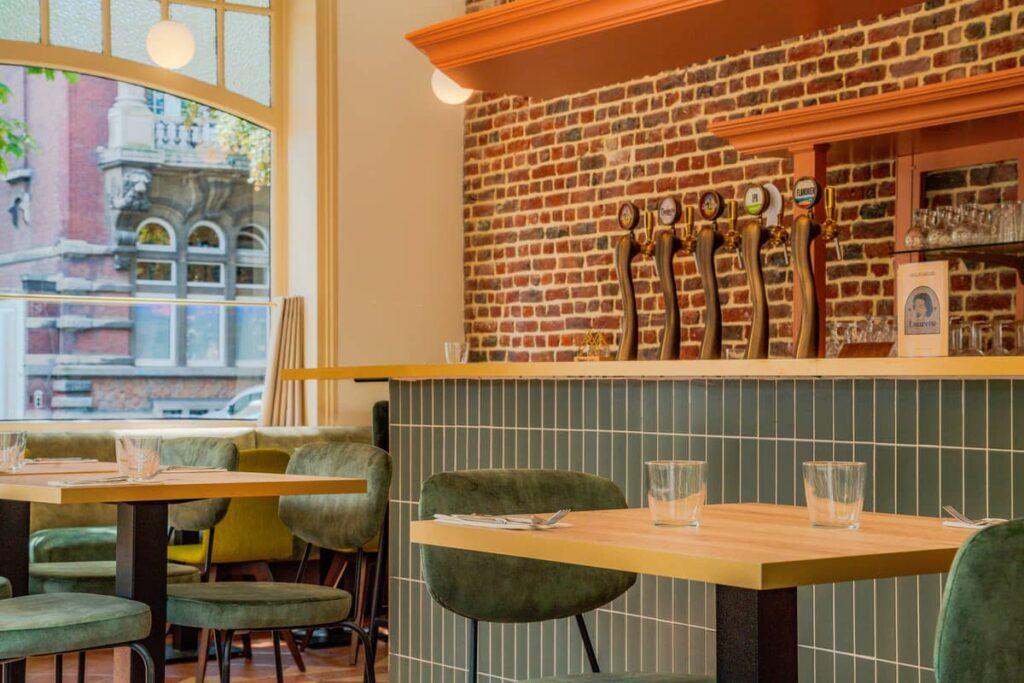 Vue du bar du restaurant Laurette, associant une faïence vert sauge posée à la verticale, des chaises en velours vert mousse, et un mur en briques apparentes qui apporte du cachet. Les luminaires suspendus et les robinets à bière dorés complètent l’ambiance chaleureuse.