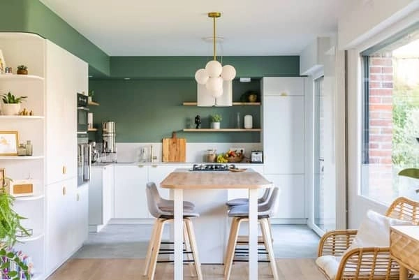 Vue d’ensemble d’une cuisine familiale aux tons doux, avec un mur vert sauge, un îlot central en bois clair avec coin repas, des meubles blancs minimalistes et des touches de laiton sur les poignées et la robinetterie.
