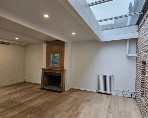 Vue d’un salon avant aménagement, avec verrière de toit, radiateur apparent et mur en briques, avant la création des meubles sur-mesure