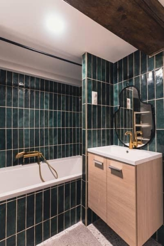 Salle de bain rénovée mêlant faïence verte, robinetterie dorée et meubles bois naturel. Architecture intérieure raffinée.